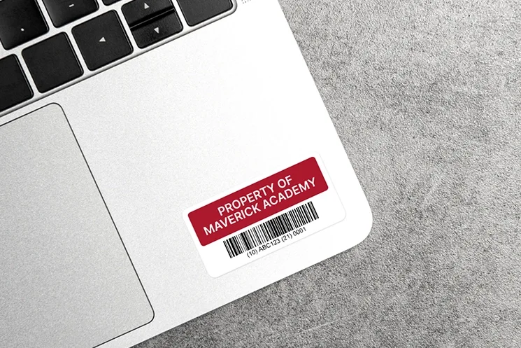 A red fixed asset label with a barcode and identification details attached to a laptop on a gray surface.