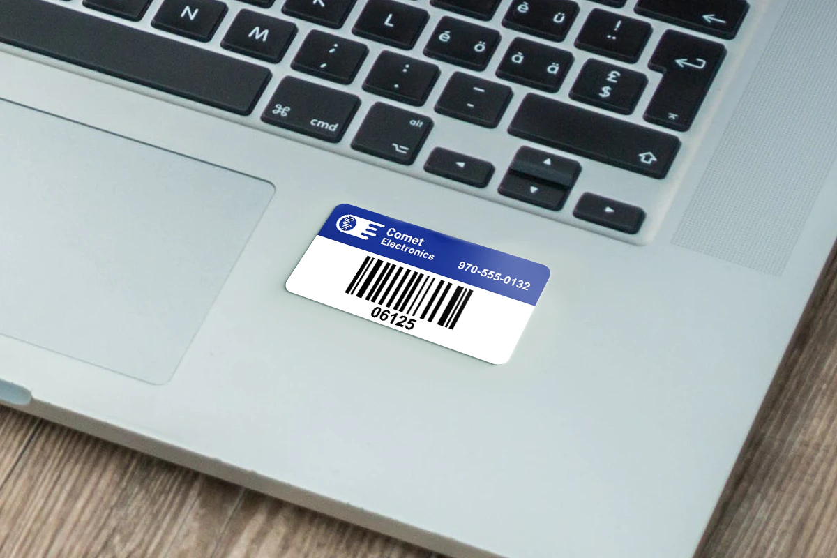 A close-up of a laptop with an asset tag sticker attached, featuring a barcode and identification details.